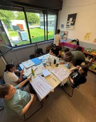 Ragazzi seduti attorno a un tavolo con fogli, colori e materiali, durante un laboratorio creativo