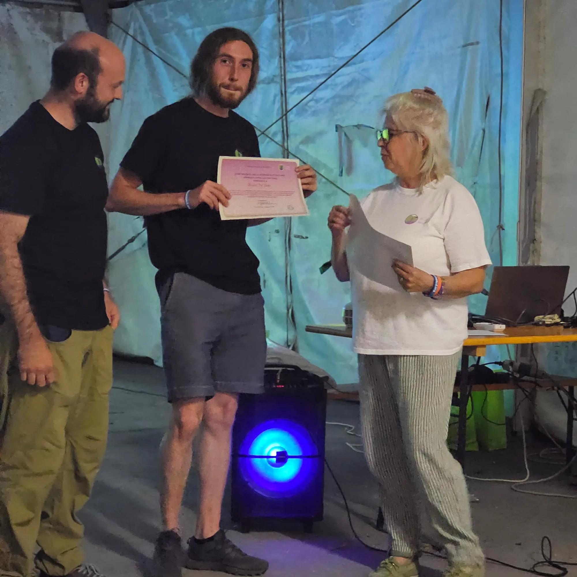 Un assegnatario mostra l'attestato della borsa di studio sotto il tendone del raduno, affiancato dall'altro assegnatario e da una rappresentante del Comitato