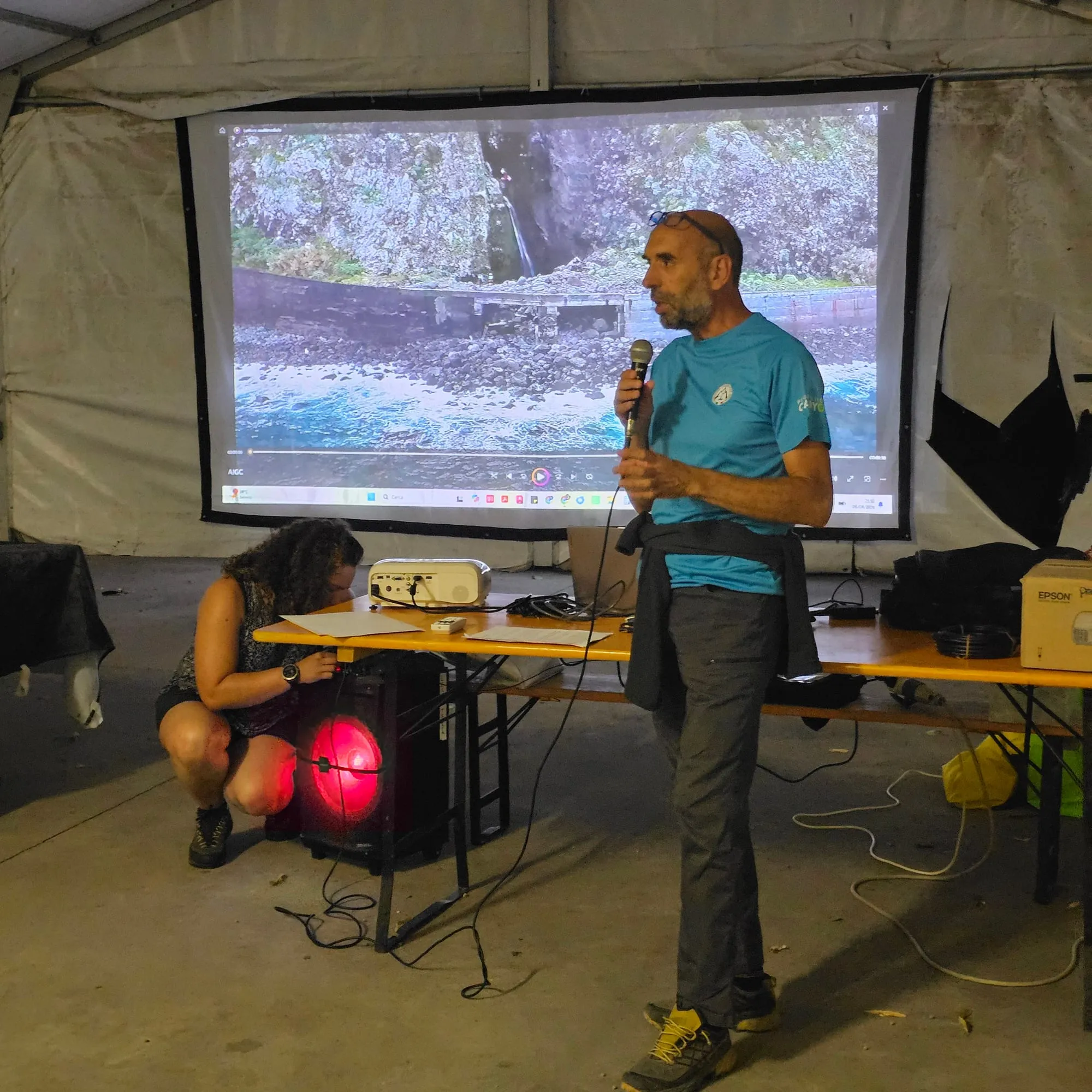Un relatore parla al microfono davanti allo schermo su cui scorre un video di canyoning, sotto il tendone del raduno