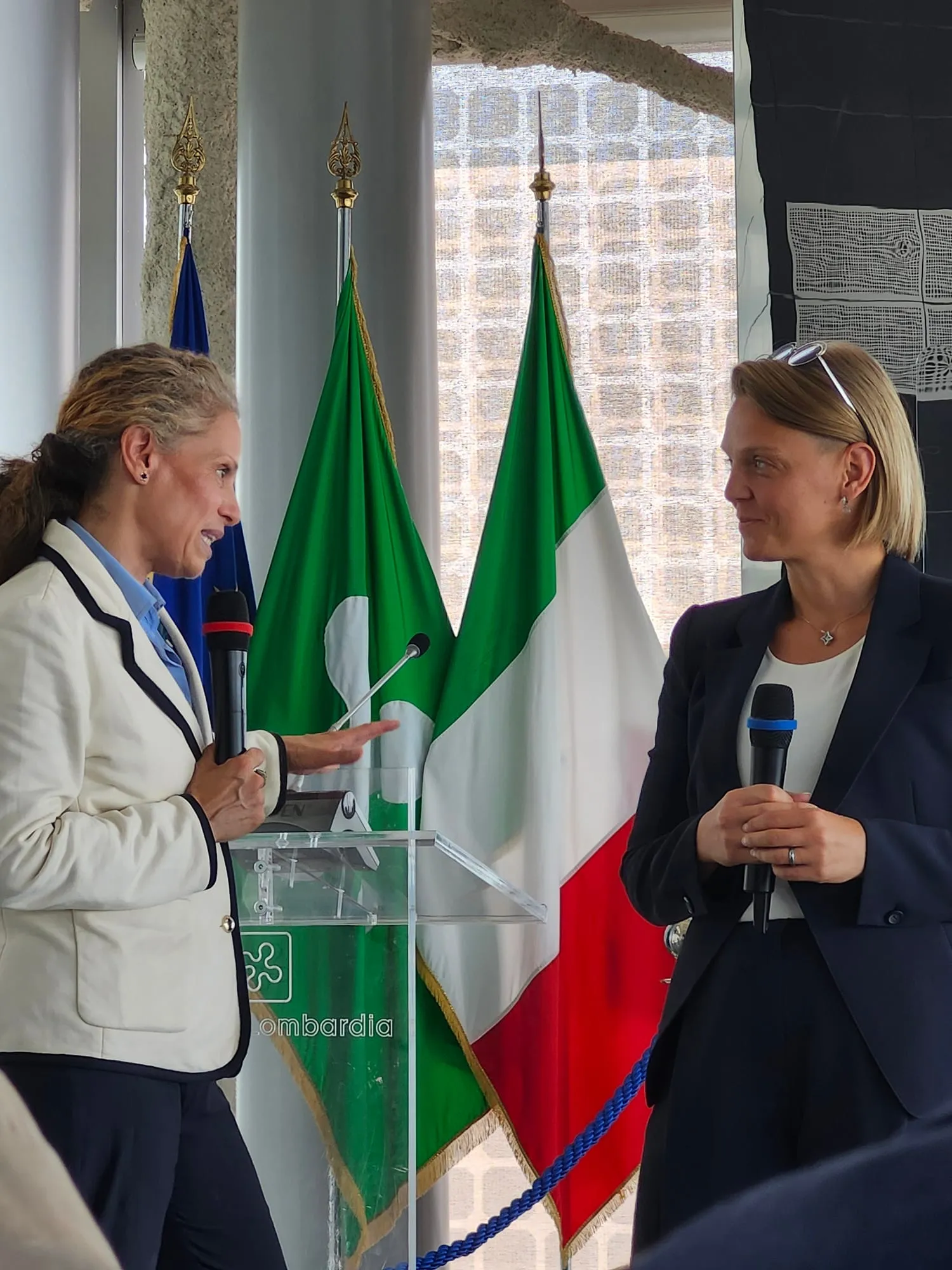 Due relatrici al microfono davanti alle bandiere europea, lombarda e italiana