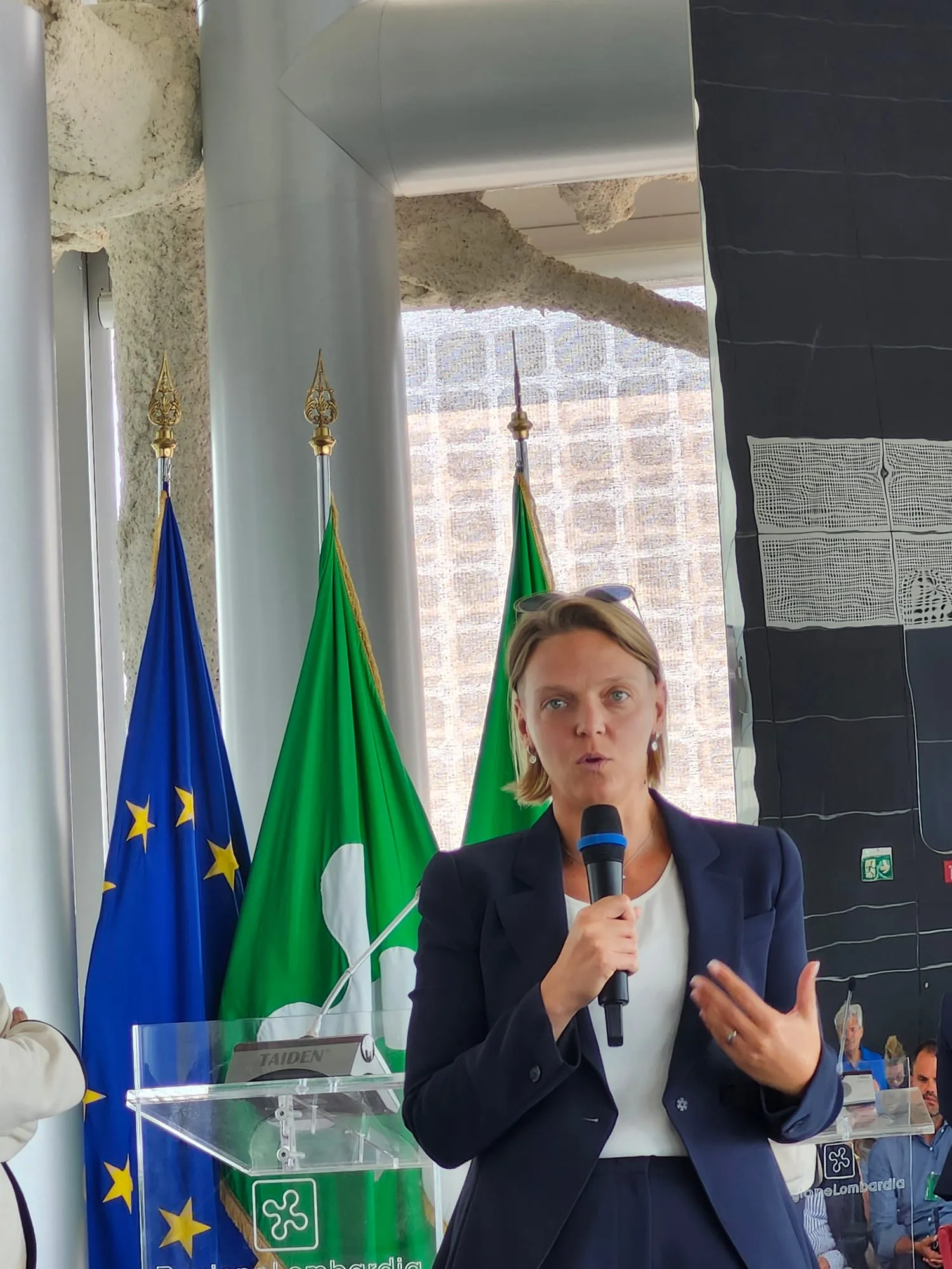 Una relatrice parla al leggio di Regione Lombardia, davanti alle bandiere