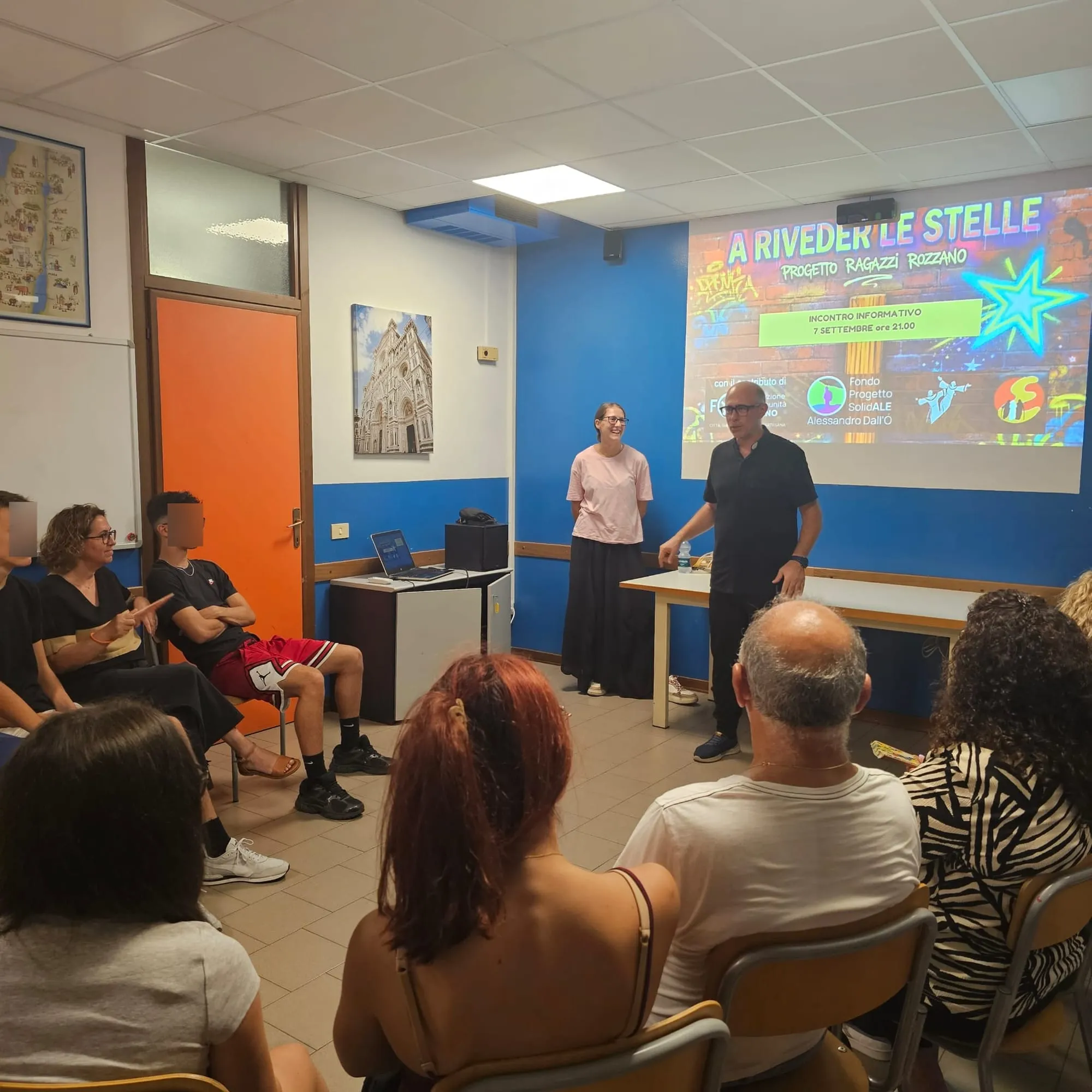 Due relatori in piedi davanti allo schermo aprono l'incontro, con il pubblico seduto attorno a loro